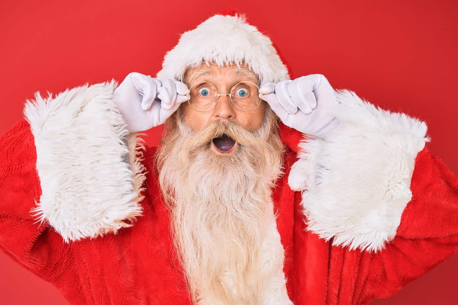 Old senior man with grey hair and long beard wearing santa claus costume holding glasses afraid and shocked with surprise and amazed expression, fear and excited face.